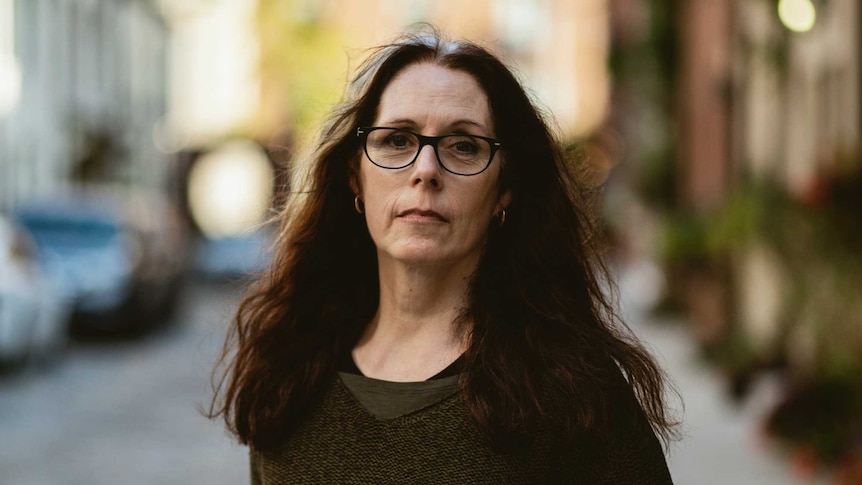 A woman with long brown hair and black thin-rimmed glasses stands with a blurred street behind her. Her expression is neutral.