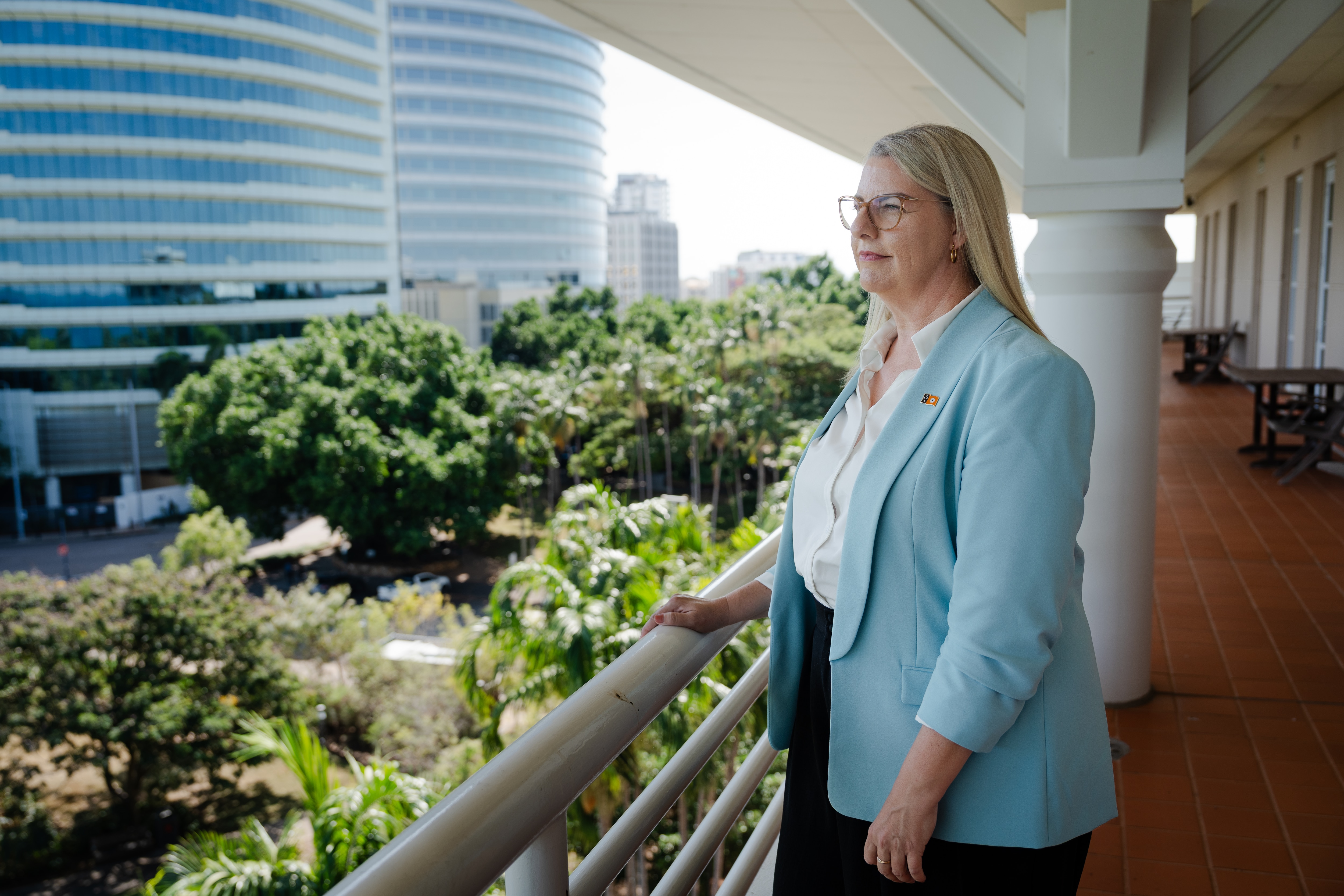 White woman, blonde long hair, in pale blue blazer, standing on Parliament building balcony, looking across Darwin landscape