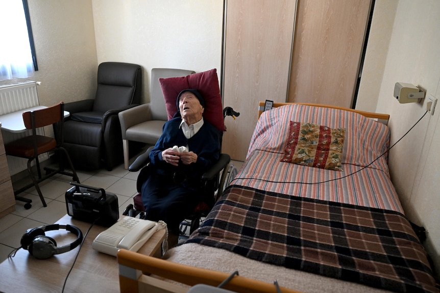 An elderly woman with her hair covered leans back in a wheelchair with a red pillow under her head in a nursing home room