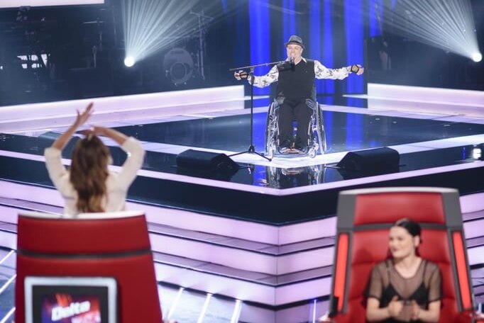 A man in a wheelchair sings on stage to a row of judges.