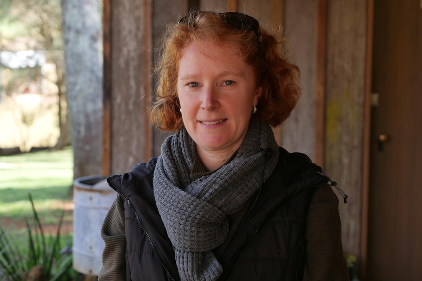 A young woman with short red hair, wearing a jacket and scarf, stands smiling outside a wooden building.