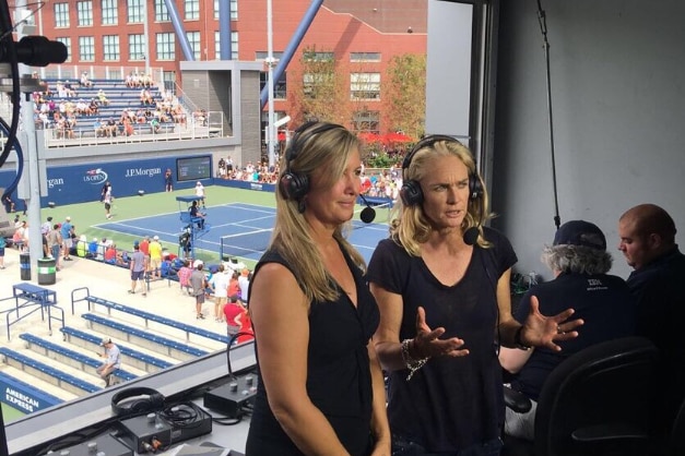 Two female commentators wear headphones in a commentary box, tennis is playing outside