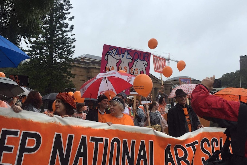 Arts supporters, holding banners and wearing orange shirts, protest on Oxford Street.