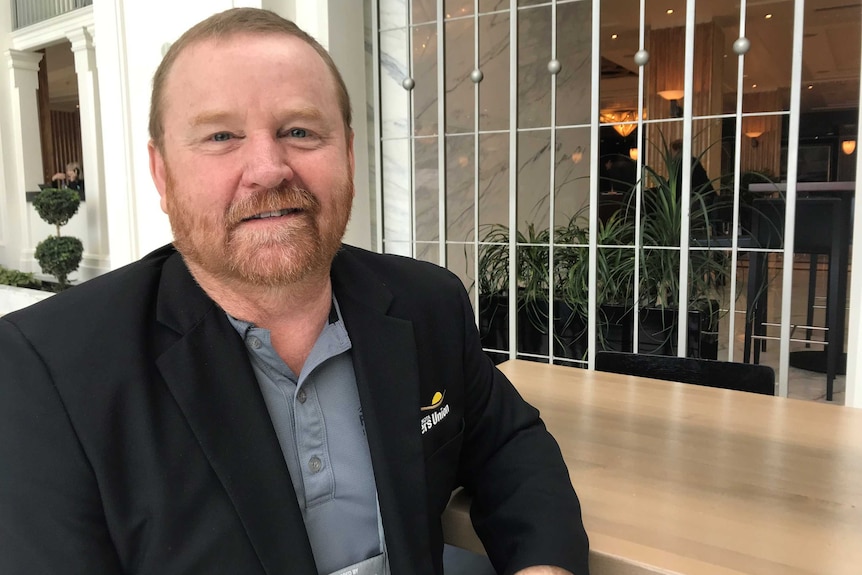 A man with short red hair and a red beard, in a black suit, seated in a hotel lobby.