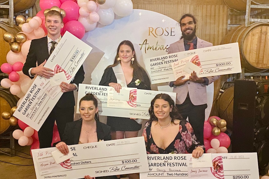 Five young people dressed up and holding large cheques in front of a backdrop of pink balloons.