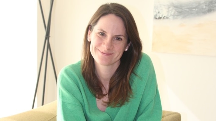 author Polly Phillips in a green jumper sitting on a chair with a pile of her latest book with an orange cover on the floor