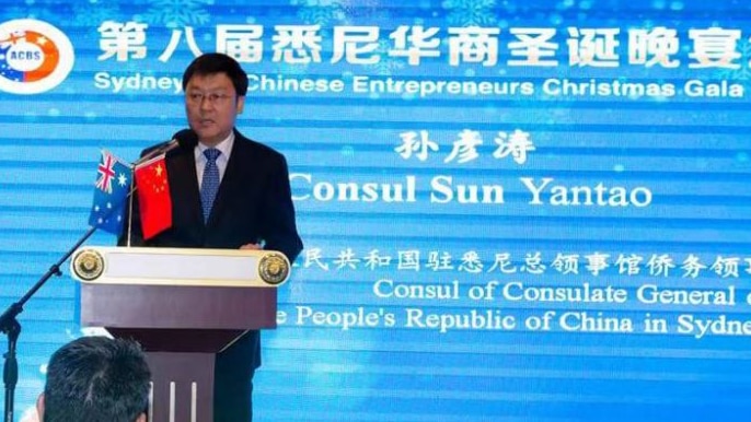 Man in suit stands on stage behind podium with large blue graphic screen behind him