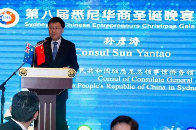 Man in suit stands on stage behind podium with large blue graphic screen behind him