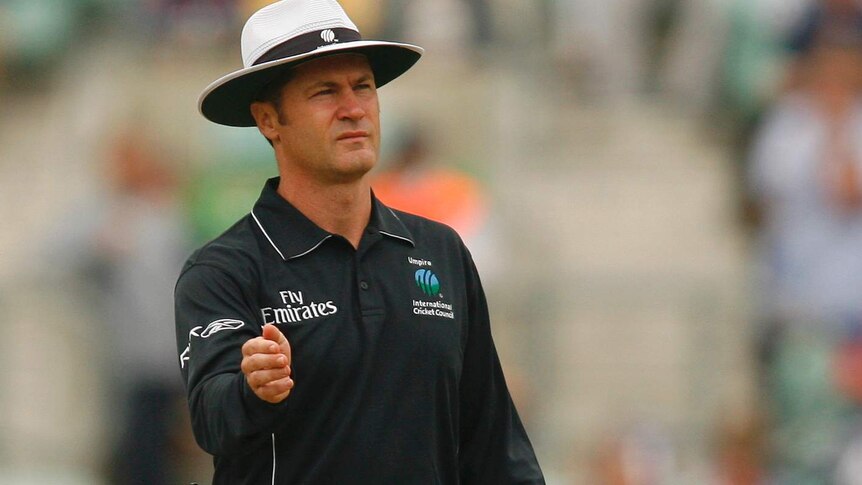 Simon Taufel is in all-black umpiring gear, holding a bowler's hat.