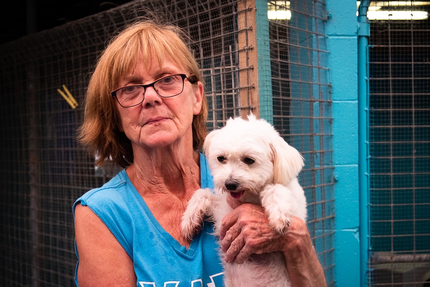 A middle-aged woman wearing a blue shirt and reading glasses holds a fluffy white dog in front of a shelter.