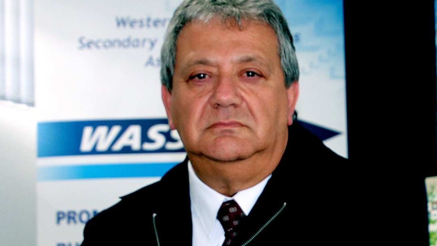 A man in a white shirt and black jacket sits at a desk in an office with a grumpy expression on his face.