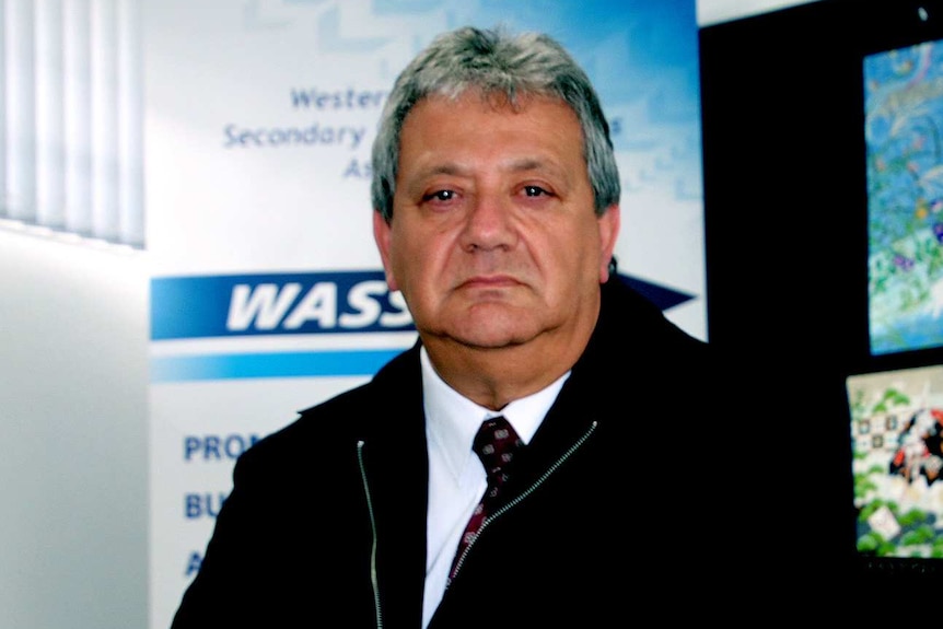 A man in a white shirt and black jacket sits at a desk in an office with a grumpy expression on his face.