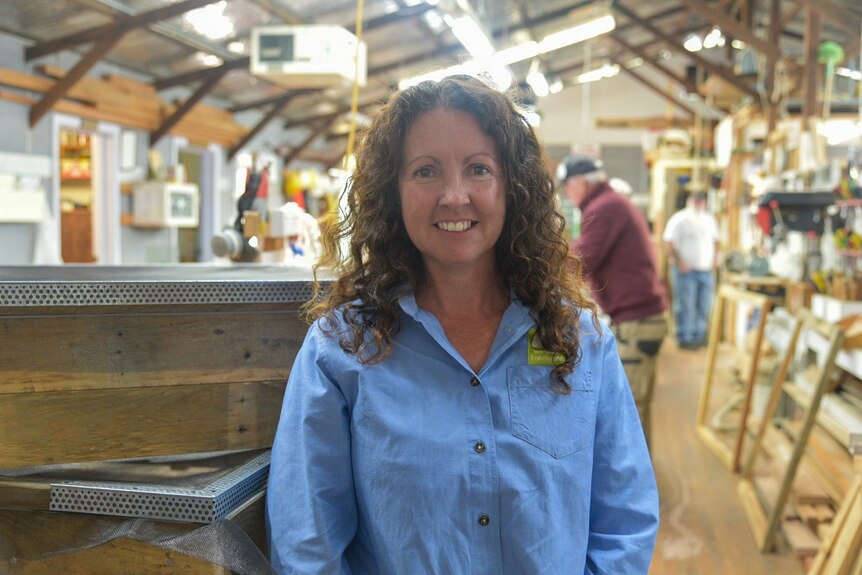 A woman with brown curly hair in a blue collared shirt stands in a cluttered workshop.