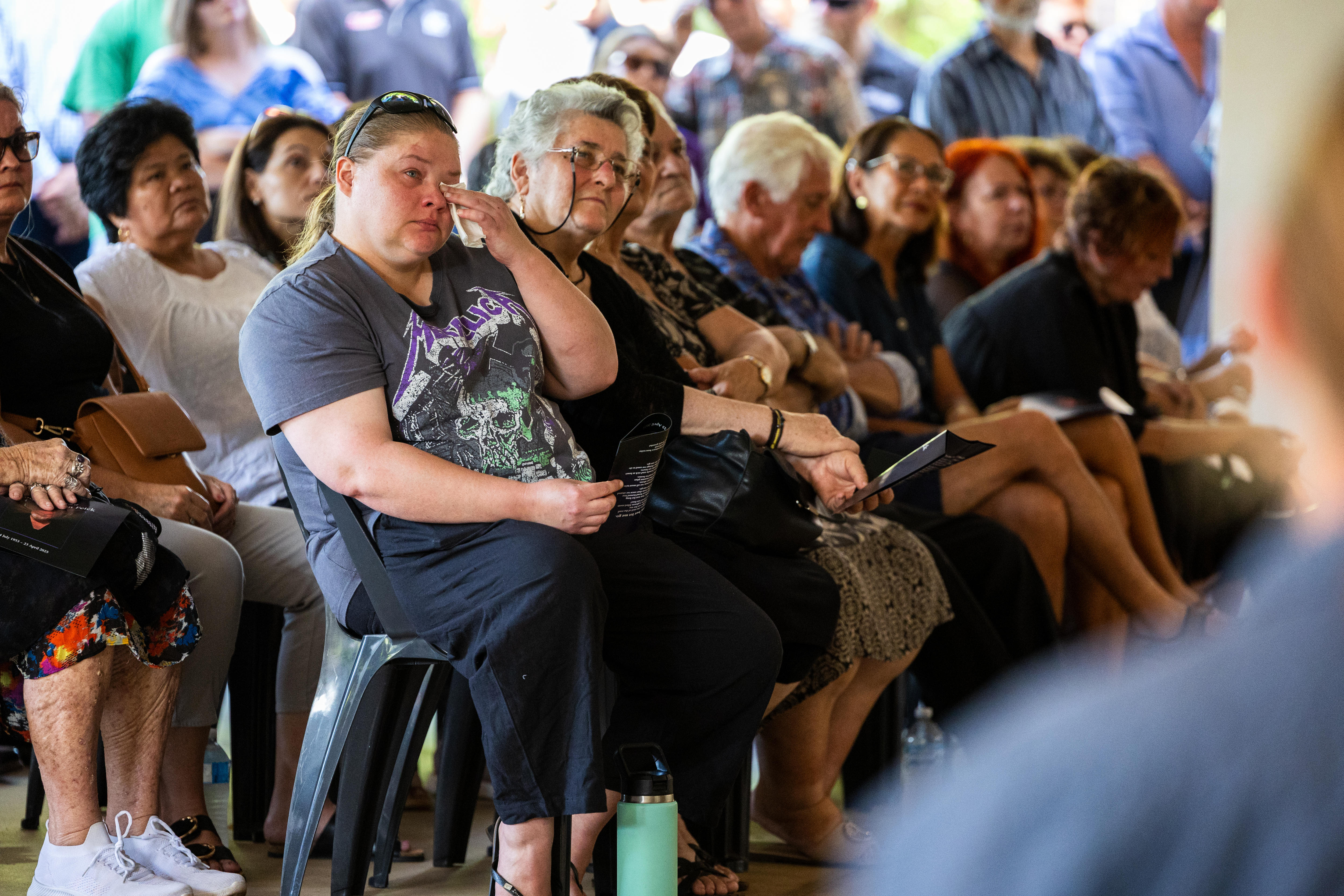 A crowd of people sitting and visibly sombre. One woman is holding a tissue to her eye, and has been crying.