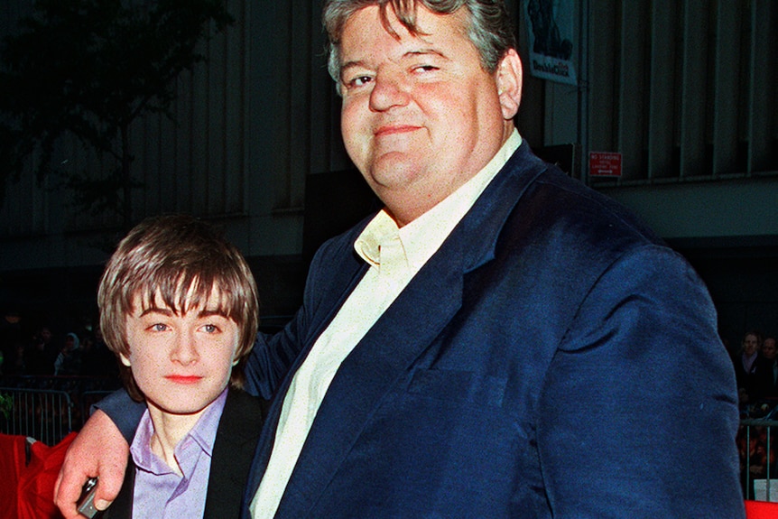 coltrane in a blue suit, white shirt on the red carpet with his arm around a young radcliff, in a quit and lavender shirt