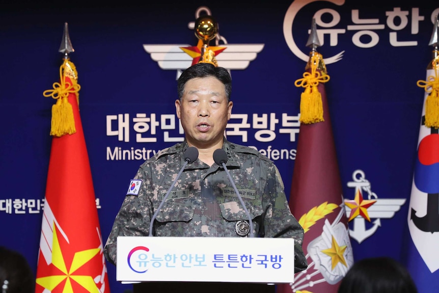 Ahn Young-ho looks straight ahead of himself as he stands in front of a blue background with flags on either side.