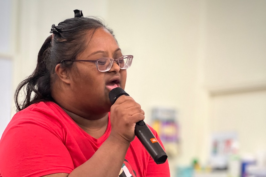 A woman in a red t shirt sings into a microphone with her eyes closed