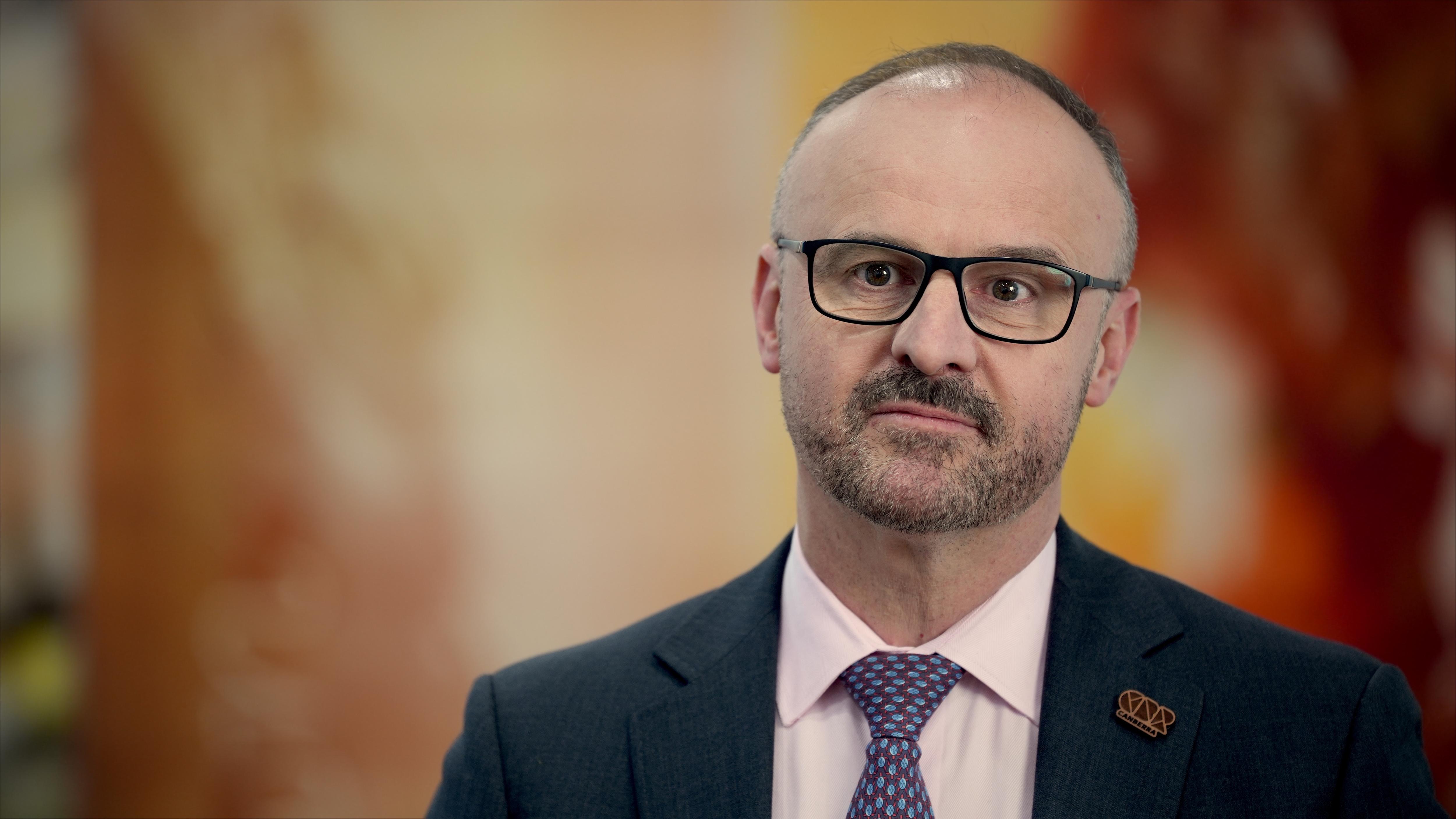 A man wearing glasses and a suit and tie stands impassively inside near artwork on the wall.
