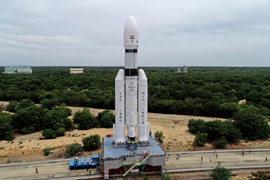 A large white space rocket sits on a railway platform, surrounded by forest with some people seen nearby.