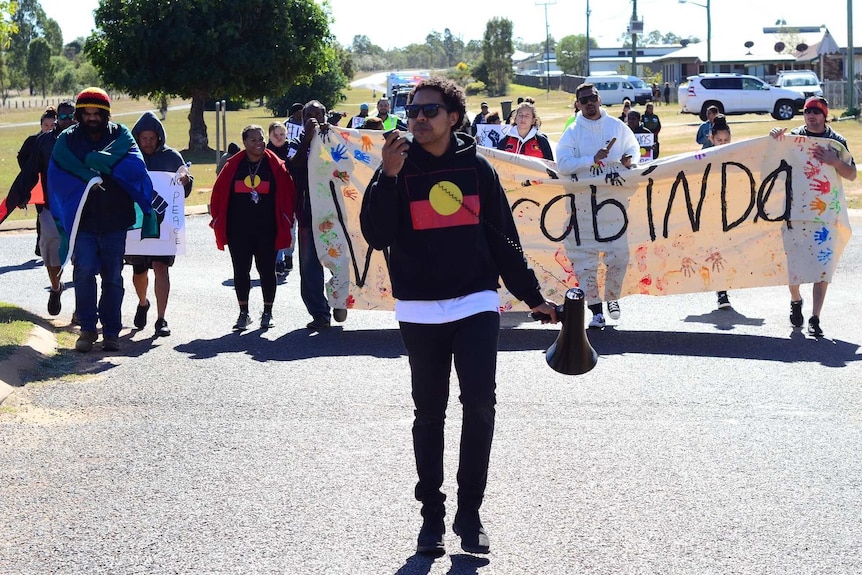 Alwyn Doolan stands with a megaphone in an Aboriginal flag hoodie in front of a crowd of people holding a Woorabinda banner.