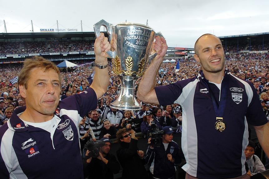An AFL coach and team captain hold the premiership cup, with thousands of fans behind them.