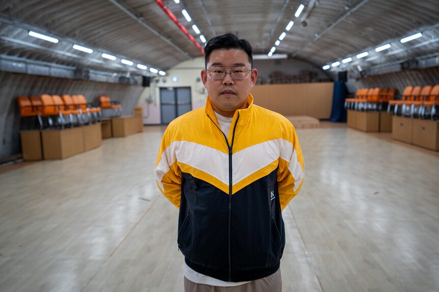 A close up of a man wearing a yellow jacket and glasses standing in a room with orange chairs stacked against the wall.