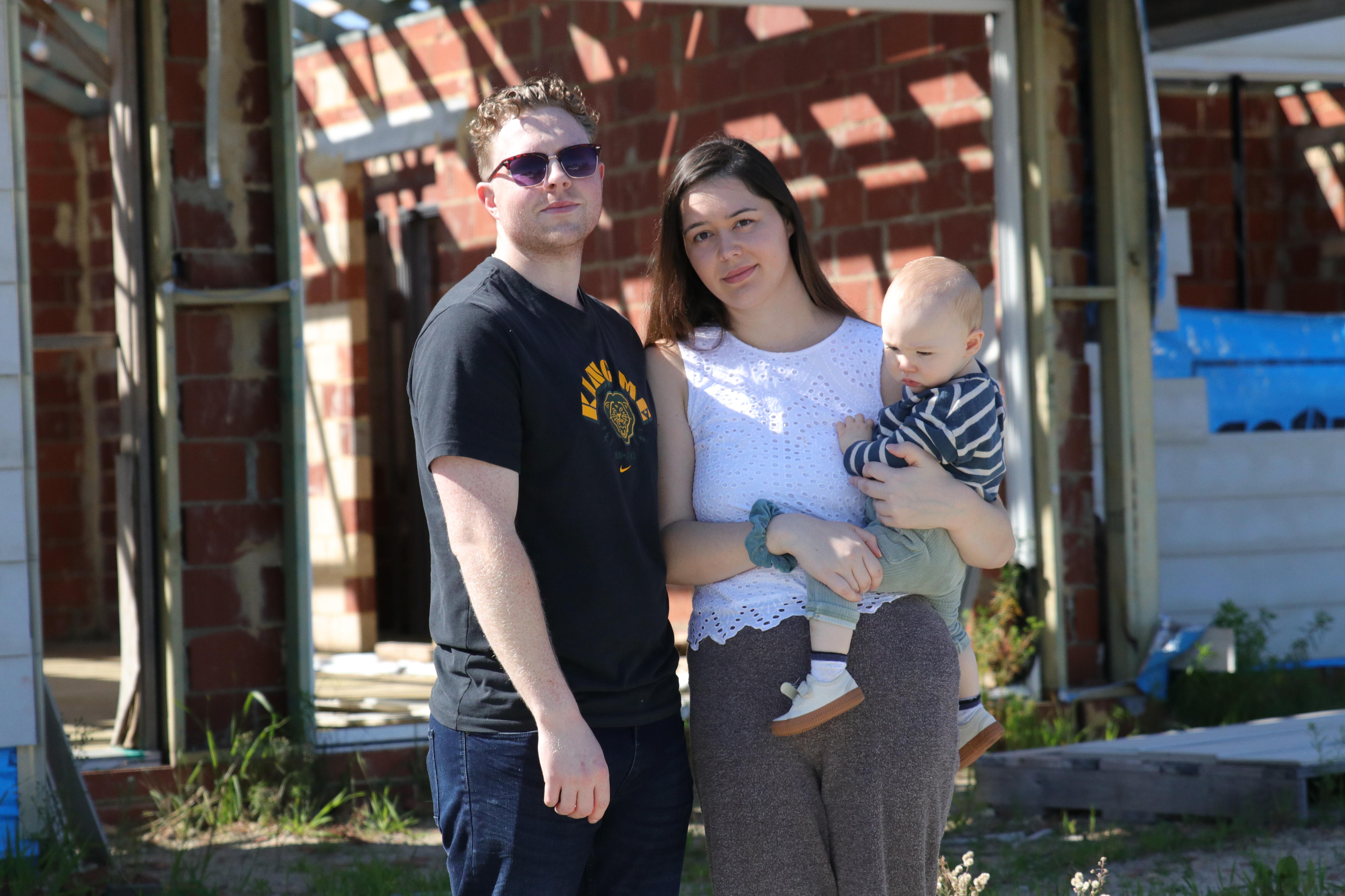 A man, woman and infant child stand in front of a house under construction.