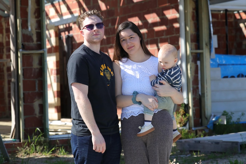 A man, woman and infant child stand in front of a house under construction.