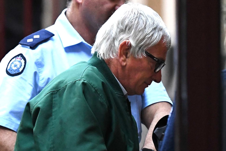 An older man with white hear, in a green jacket, is escorted into court by prison guards.