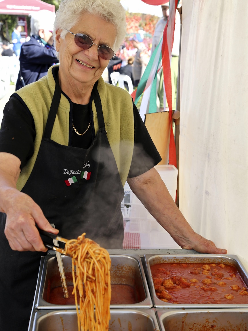 A woman in an apron holding a pair of tongs serving pasta 
