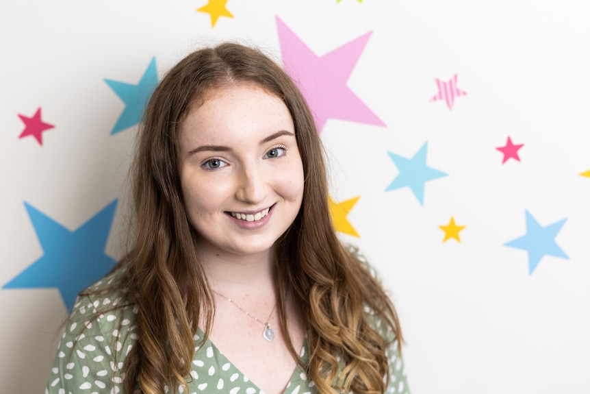 A young woman in front of a colourfully decorated wall smiles at the camera.