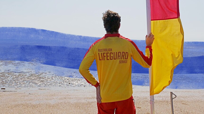 A surf lifesaver stands on the beach holding a red and yellow flag looking out over the ocean.