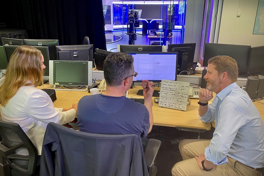 Three people sit at a computer chatting, with a TV studio lit up in the background