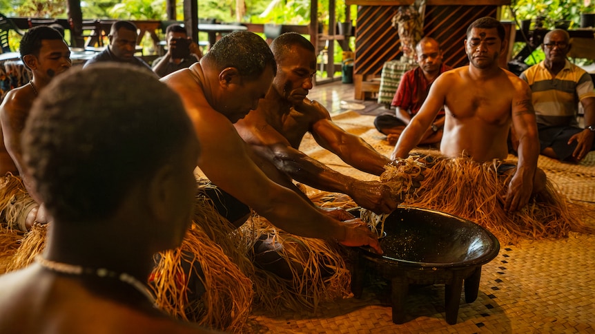 Photo of multiple men in ritual clothing making a drink out of roots.