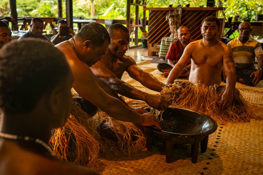 Shirtless men in grass skirts sit around a large bowl, mixing up a concoction.