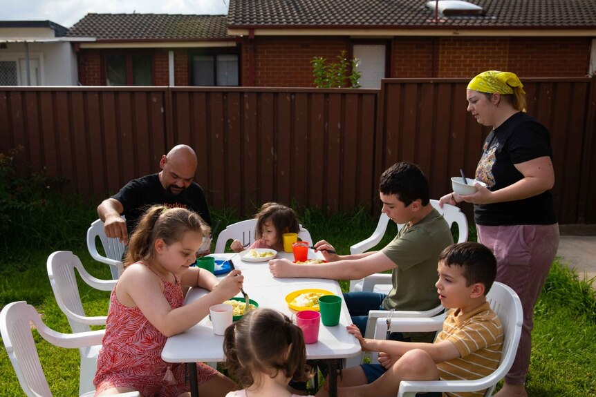 A group of people eat at an outdoor white table in a backyard.