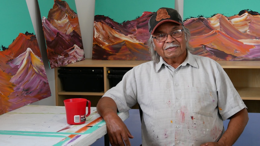 A man wearing a cap and glasses sits on a chair in front of painted landscapes of mountains.