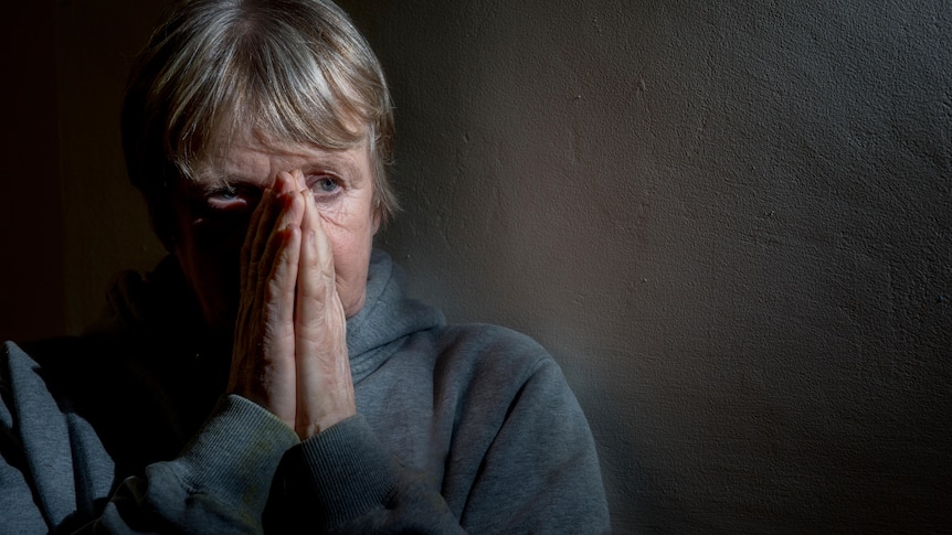 older woman places hands together in front of her face looking sadly into the distance against black background