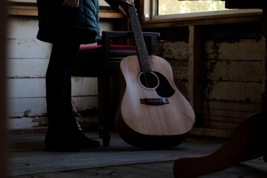 A dirty guitar leaning on a chair inside a gutted house with a woman standing next to it