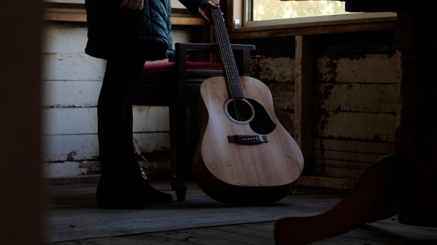 A dirty guitar leaning on a chair inside a gutted house with a woman standing next to it