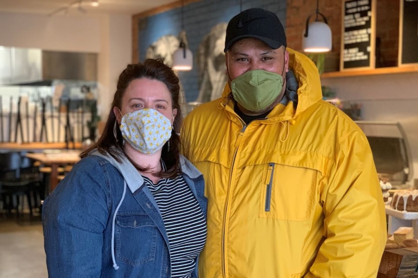 A woman in a denim jacket and a man in a black cap and yellow both wear face masks and stand in an empty cafe.