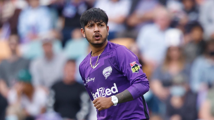 A young Nepalese man wearing a purple BBL uniform as well as a gold chain and white watch looks intense on a cricket field.