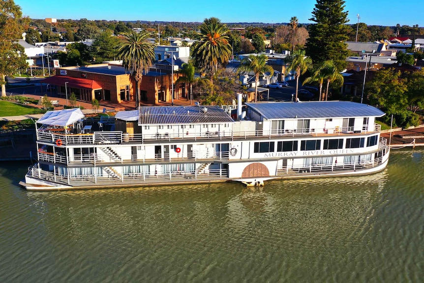 A large paddleboat moored on the a wide body of water with buildings and trees in background. 