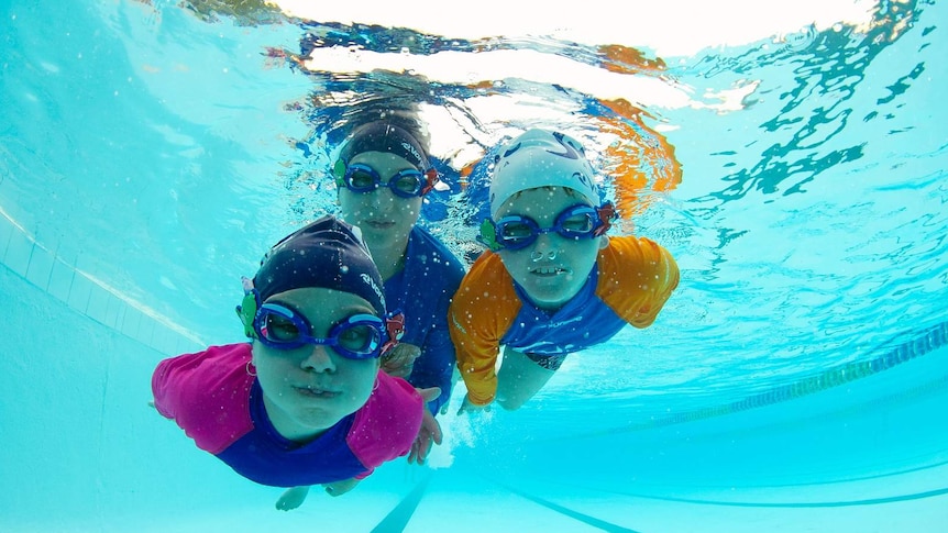 Three kids swim towards the camera below the surface of a pool, lanes visible behind them. They wear goggles and swimming caps.