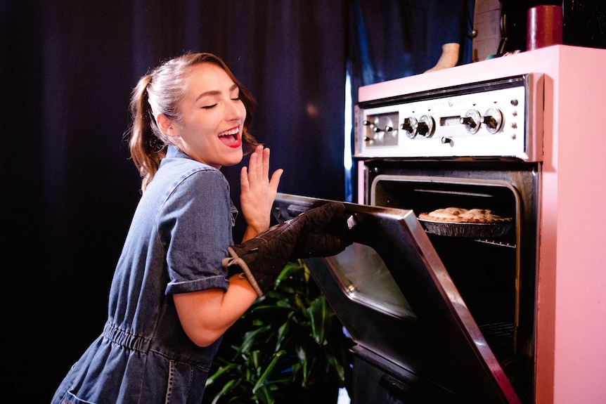 A woman in a blue dress and red lipstick smiles as she kneels at a pink retro style oven