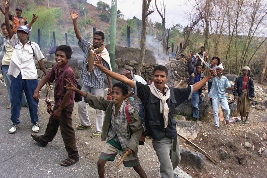 A group of men and boys wave their hands and smile from the side of a road. Behind them smoke from a fire rises.