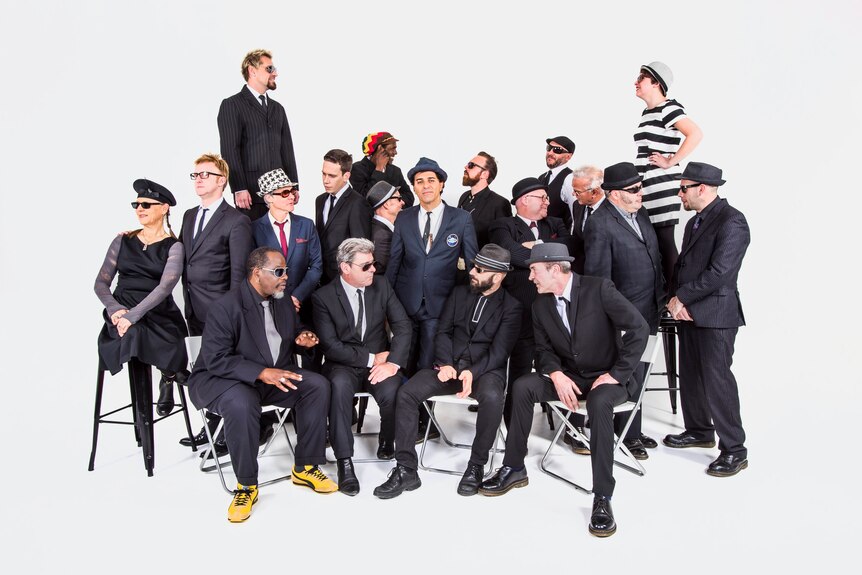 A group of about 20 people in interesting poses wearing dark suits, hats and sunglasses in a white room.
