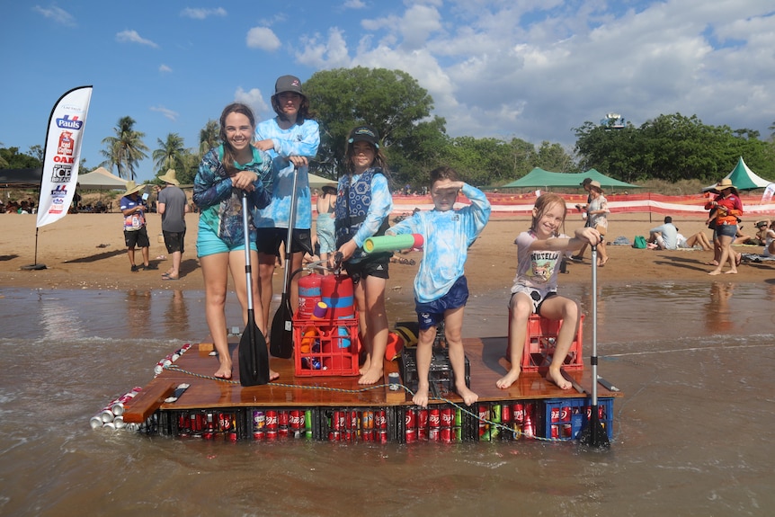 Five children in brightly coloured clothes and bathers standing on a boat made out of soft drink cans, on a beach. 