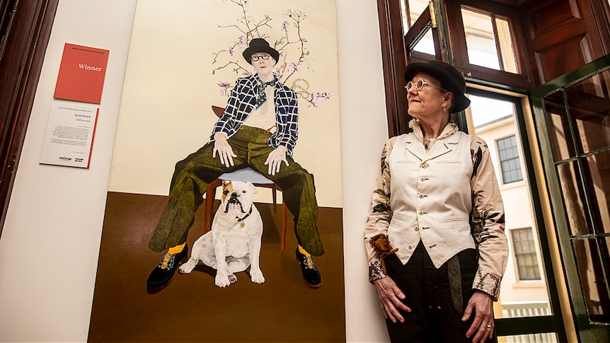 Woman with short hair and dark brown felt hat looking at her own self-portrait on the wall. Wearing white button-down vest.