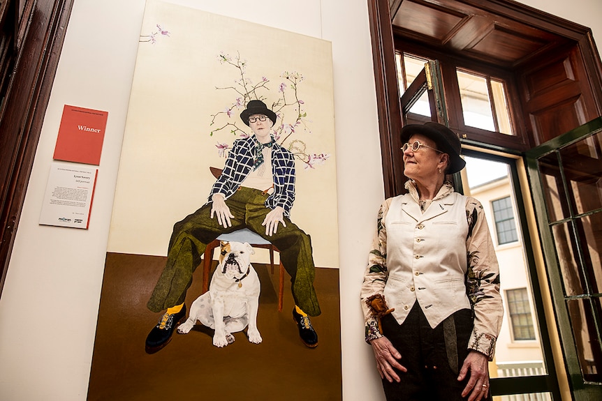 Woman with short hair and dark brown felt hat looking at her own self-portrait on the wall. Wearing white button-down vest.